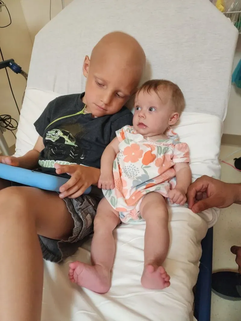 طفل مريض بمرض السرطان يجلس على سرير مستشفى مع شقيقته الرضيعة، حيث يظهر تفاعل حنون بينهما وسط الأجواء الطبية.