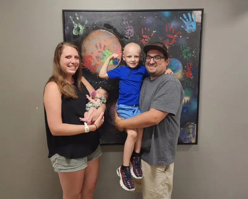 عائلة فيلا تتوسط الصورة، حيث يحمل الأب كودي ابنه كايلين المصاب بالسرطان، بينما تحمل الأم آلي ابنتهم حديثة الولادة، مع خلفية فنية ملونة.