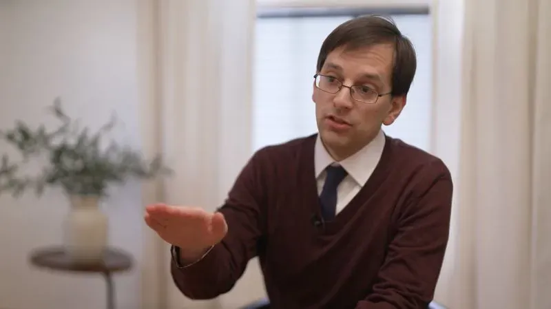 شخص يتحدث بجدية في غرفة مضاءة جيدًا، مع التركيز على قضايا علاج السرطان وأهمية التمويل للبحوث الطبية.