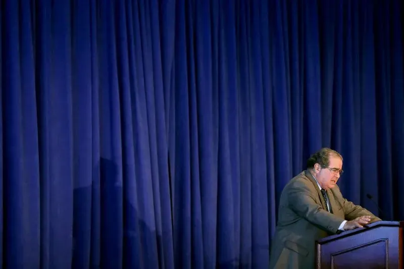 صورة لقاضي المحكمة العليا الراحل أنتونين سكاليا يتحدث أمام جمهور، مع ستارة زرقاء خلفه، تعكس أسلوبه الجريء في النقاشات القانونية.