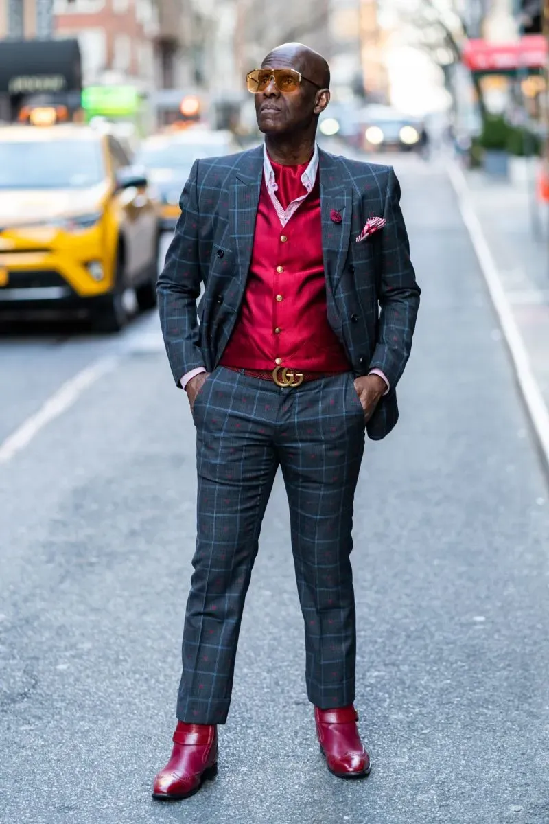رجل يرتدي بدلة زوت أنيقة مزينة بنقوش، مع قميص أحمر وحذاء أحمر لامع، يتجول في شارع حضري، يعكس أسلوب الداندية في الموضة.