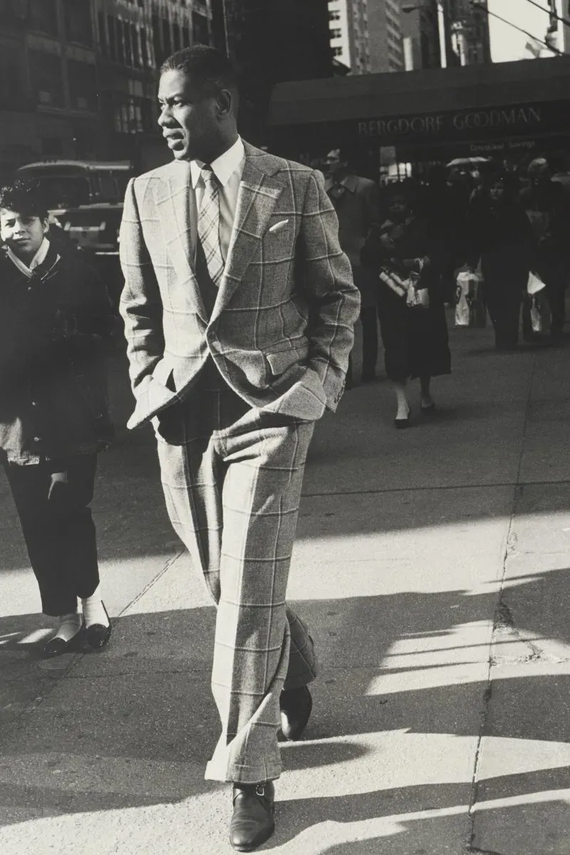 رجل يرتدي بدلة زوت مخططة، يسير في شارع حضري مزدحم، مع أطفال ومارة في الخلفية، يعكس أناقة عصر نهضة هارلم وتأثير الموضة السوداء.