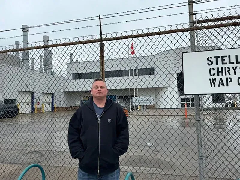 رجل يقف أمام مصنع ستيلانتيس، مع لافتة تشير إلى اسم المصنع، في كوكومو بولاية إنديانا، حيث يتأثر العمال بتعريفات السيارات.