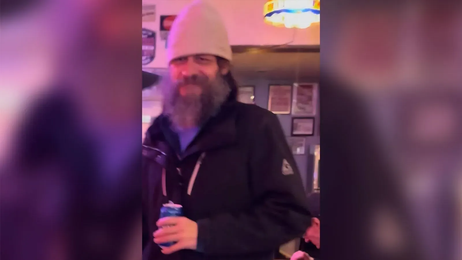 صورة لرجل ذو لحية وشعر طويل يرتدي قبعة بيضاء ومعطف داكن، يحمل علبة مشروب في حانة. يُشتبه فيه بارتكاب جريمة قتل في مونتانا.