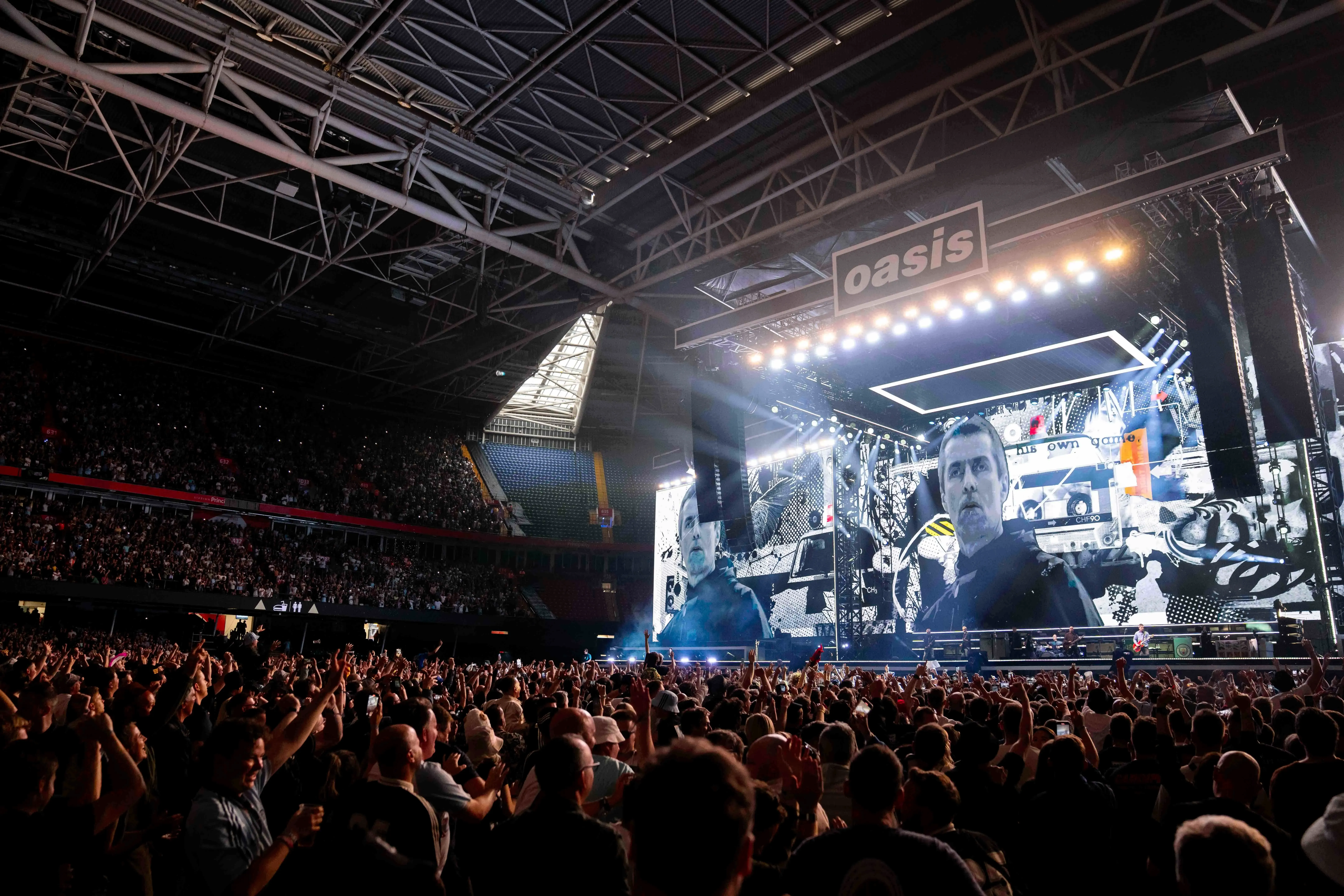 حفل فرقة Oasis في استاد ويمبلي، حيث crowd كبير يتفاعل مع الأداء، وسط أجواء حماسية بعد حادث مأساوي.