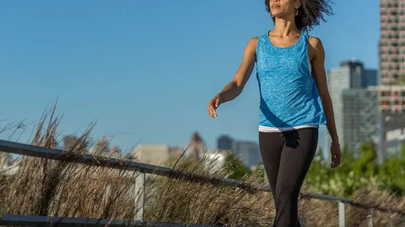 امرأة تمارس المشي في منطقة حضرية، ترتدي قميصًا أزرق وسروالًا أسود، وسط طبيعة خضراء ومباني خلفية، تعكس أهمية النشاط البدني أثناء التعافي من المرض.