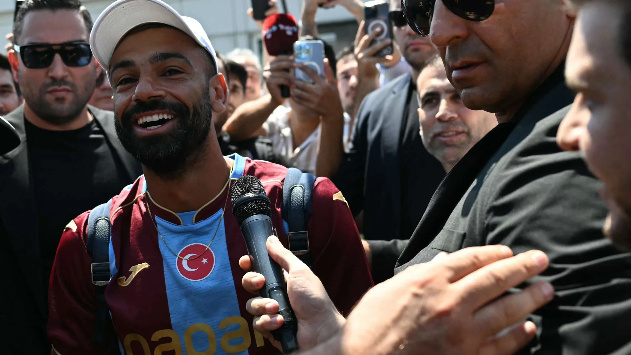 محمد صلاح يرتدي قميص Trabzonspor ويبتسم وسط حشد من الصحفيين والمشجعين خلال وصوله إلى تركيا لبدء فصل جديد في مسيرته الاحترافية.