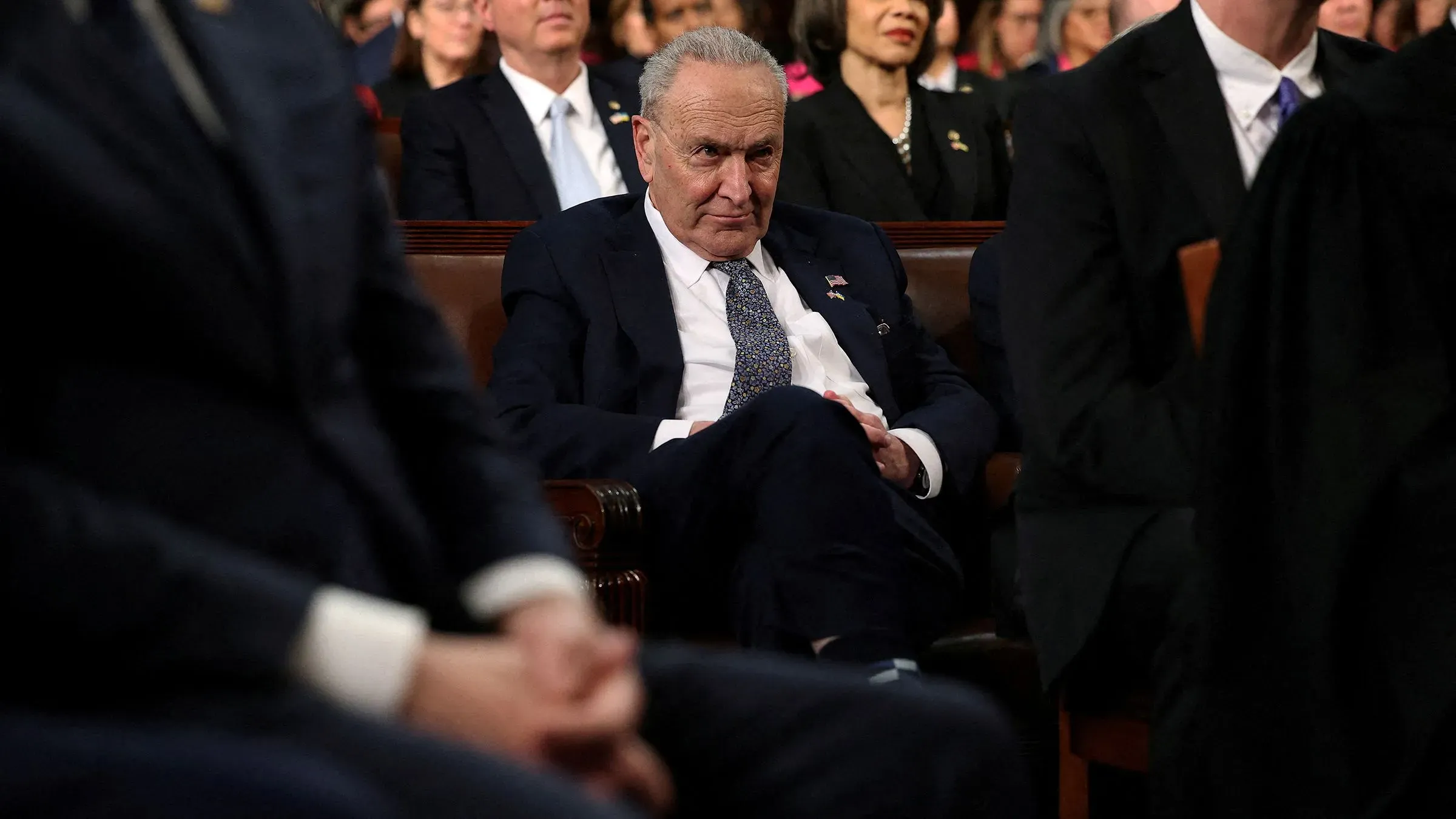 تشاك شومر، زعيم الديمقراطيين في مجلس الشيوخ، يجلس بتعبير جاد خلال حدث رسمي، مع أعضاء آخرين في الخلفية، مما يعكس ضغوط السياسة الحالية.