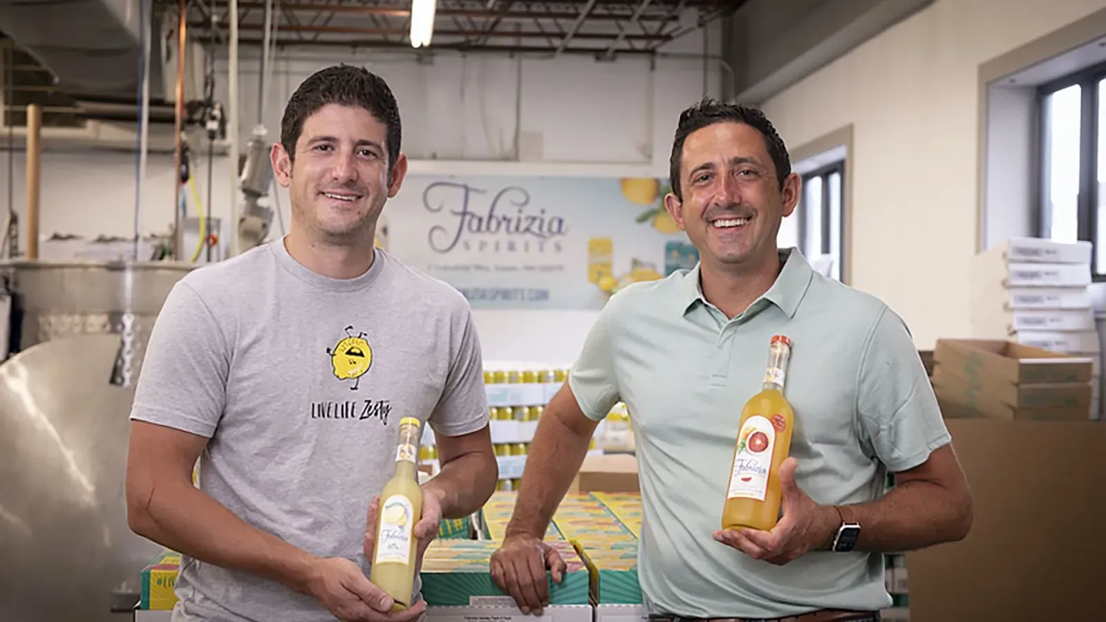 رجلان يقفان في مصنع "فابريزيا سبيريتس"، يحمل كل منهما زجاجة مشروب ليمون، مع خلفية تظهر منتجاتهم.
