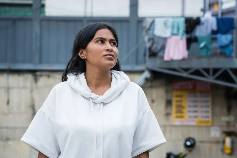 امرأة شابة ترتدي سترة بيضاء، تنظر إلى السماء في فوساغاسوغا، مع خلفية تضم ملابس معلقة، تعكس تحديات حياة ما بعد السجن.