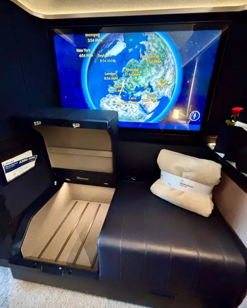 مقصورة درجة أولى فاخرة على متن طائرة، مع شاشة عرض توضح مسار الرحلة، ومقاعد مريحة مزودة بوسائد، تعكس تجربة سفر متميزة.