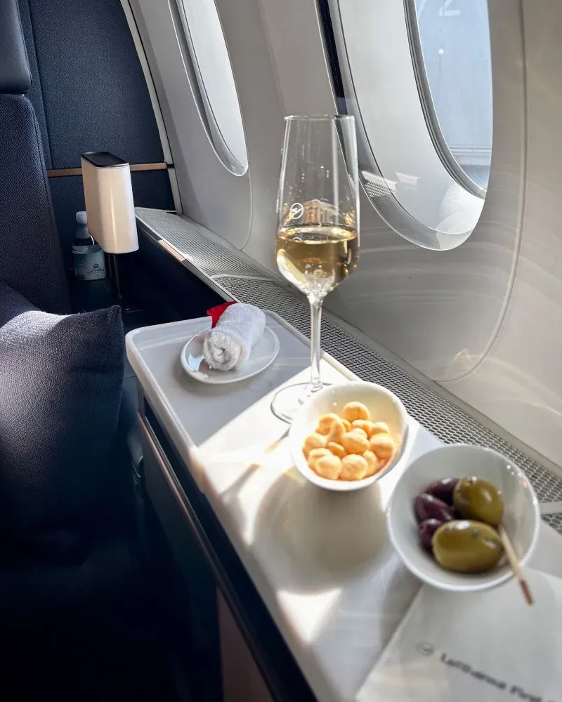 مشهد داخلي لدرجة الأولى في طائرة لوفتهانزا، يظهر كأس شمبانيا ومقبلات بجانب نافذة، مما يعكس تجربة سفر فاخرة ومريحة.