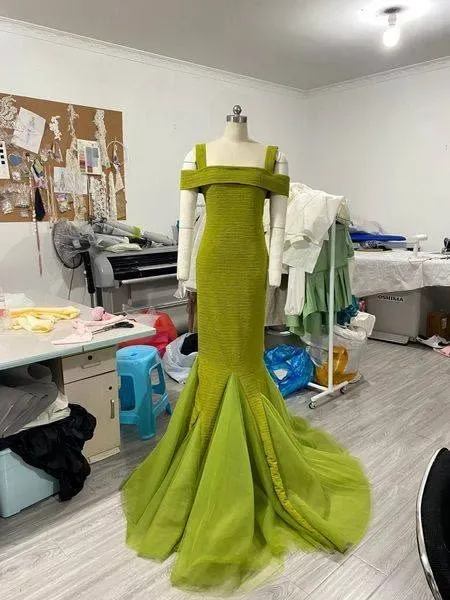 فستان طويل بلون أخضر زاهي مع تصميم أنيق، معروض على عارضة في ورشة عمل. يظهر خلفية تحتوي على أدوات خياطة ومعدات.