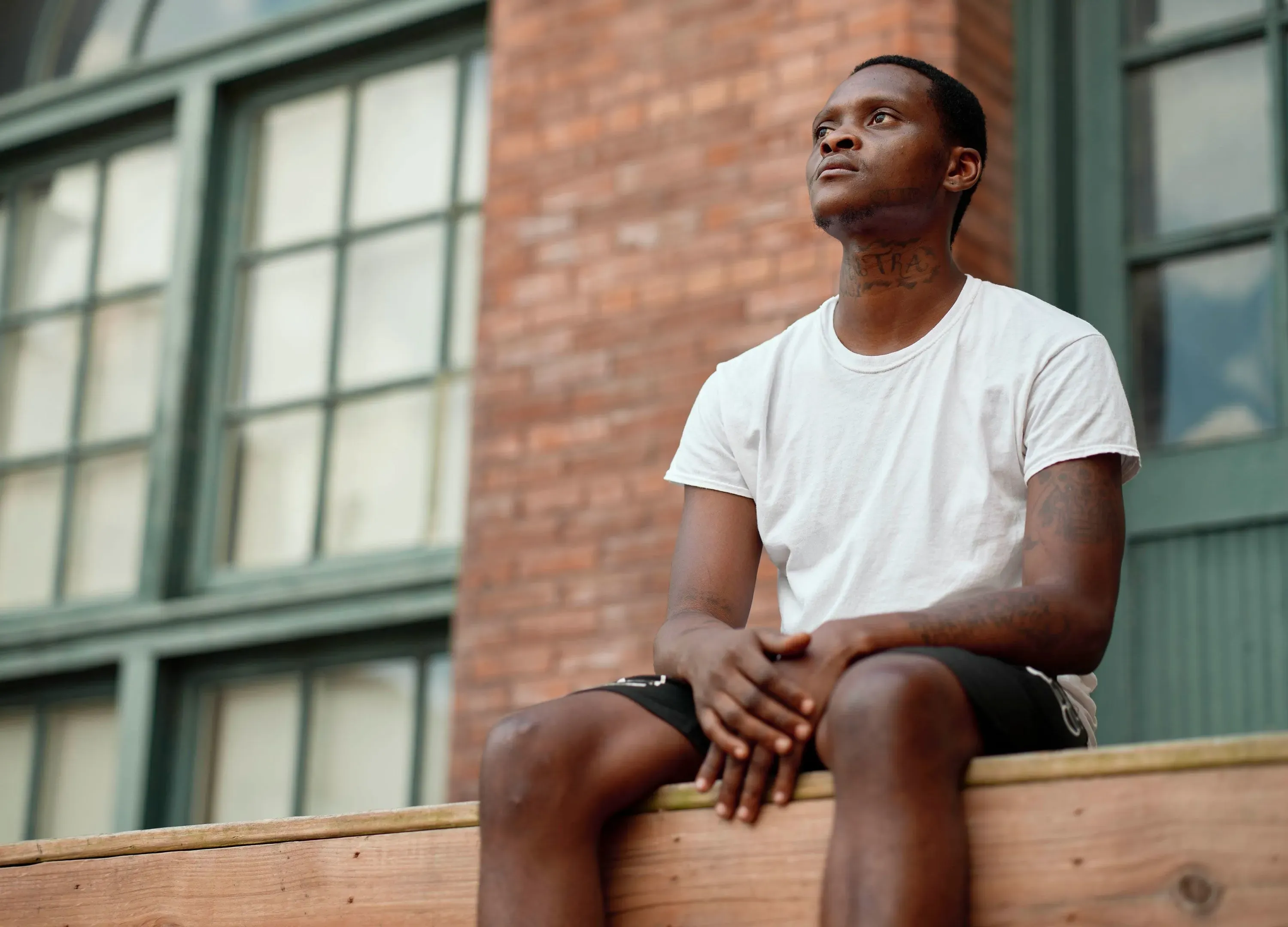 شاب يجلس على حافة منصة خشبية، ينظر بتفكير إلى الأعلى، مع خلفية من نوافذ زجاجية كبيرة، يعكس رحلة الشفاء بعد إصابة خطيرة.