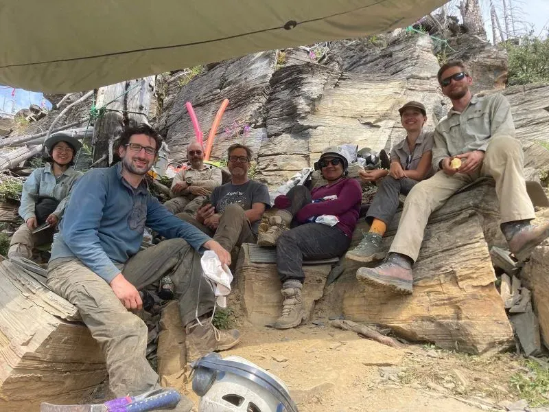 مجموعة من علماء الحفريات يجلسون في موقع حفر، محاطين بالصخور والنباتات، أثناء بحثهم عن بقايا حيوانات بحرية قديمة.