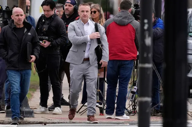 رجل يرتدي بدلة رمادية يسير نحو المحكمة، محاطًا بمصورين وصحفيين، في سياق إعادة محاكمة كارين ريد بتهمة القتل.