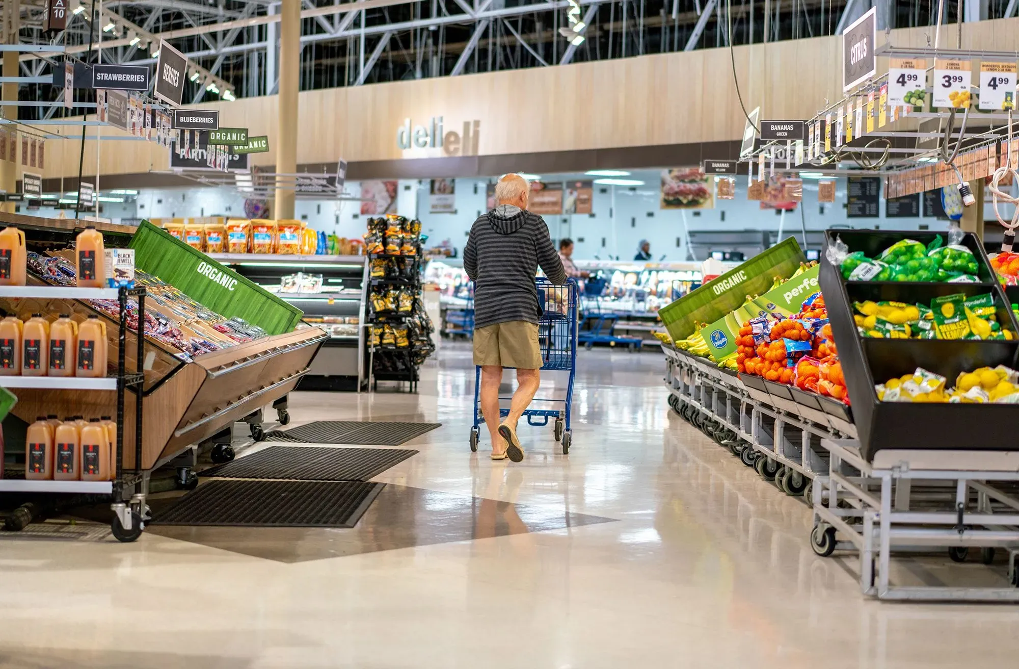 رجل مسن يتسوق في سوبرماركت، محاط بأرفف مليئة بالفواكه والخضروات، مع تركيز على ارتفاع أسعار المواد الغذائية.