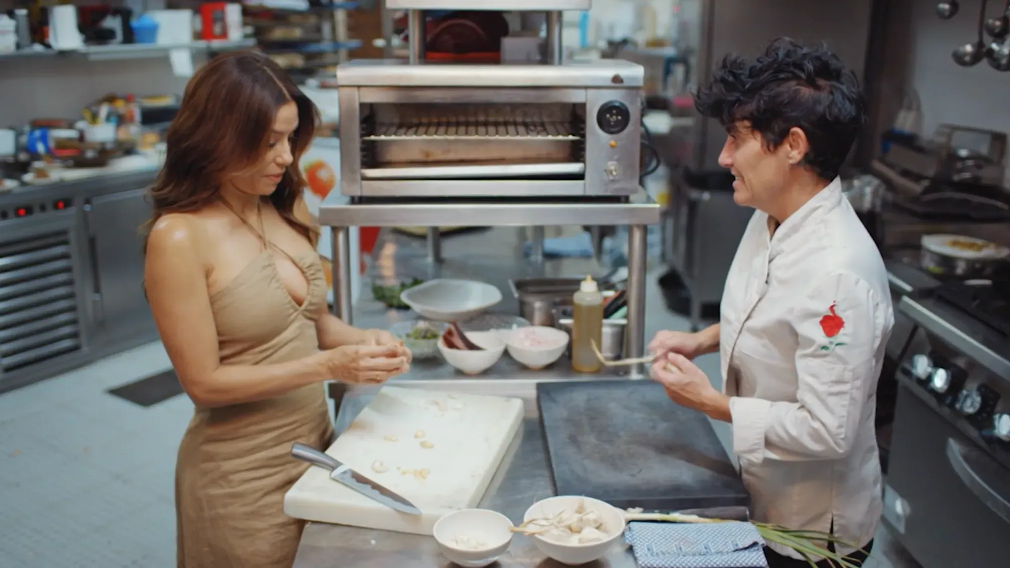 شيفتان تعملان في مطبخ، واحدة تقوم بتقشير الثوم بينما الأخرى تحمل ملعقة، مع أدوات ومكونات الطهي في الخلفية.