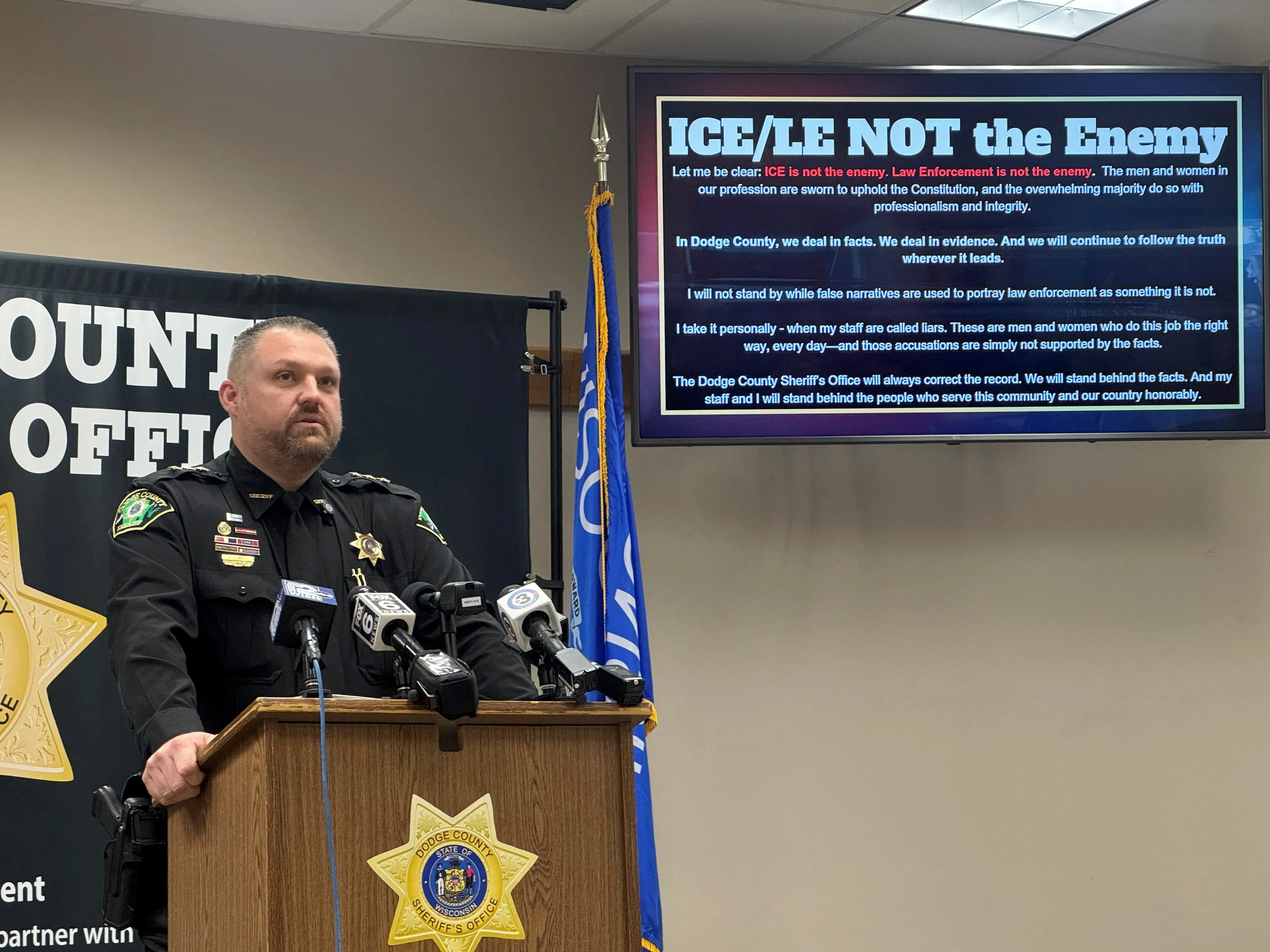 شريف مقاطعة Dodge يتحدث في مؤتمر صحفي، مع شاشة تعرض بيانًا يؤكد عدم صحة ادعاءات احتجاز Sundas Naqvi من قبل ICE.