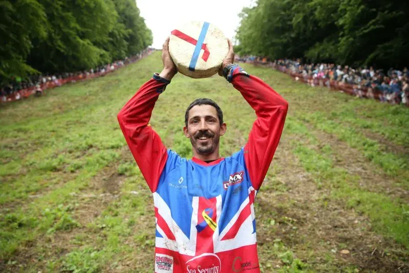 فائز يحمل عجلة جبن جلوستر بعد سباق دحرجة الجبن في بروكوورث، وسط حشد من المتفرجين في الخلفية.