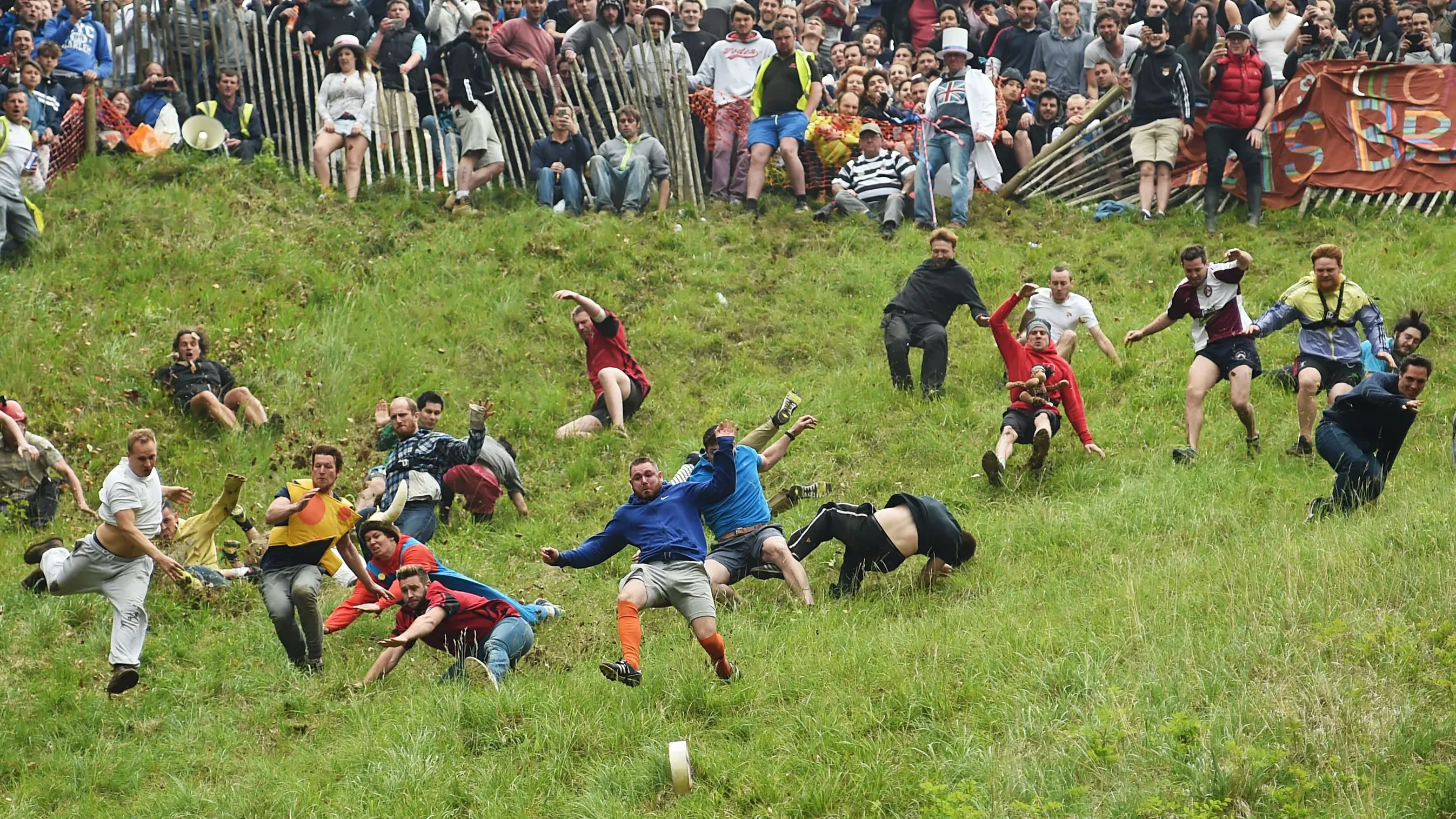 متسابقون يتدحرجون على تل شديد الانحدار خلال سباق دحرجة الجبن في بروكوورث، مع حشد كبير يشاهد من الخلف.