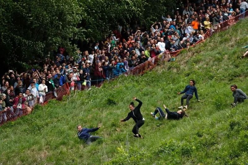 متسابقون يتدحرجون على تلة شديدة الانحدار خلال سباق دحرجة الجبن، بينما يتجمع حشد كبير من المشاهدين في الخلفية.