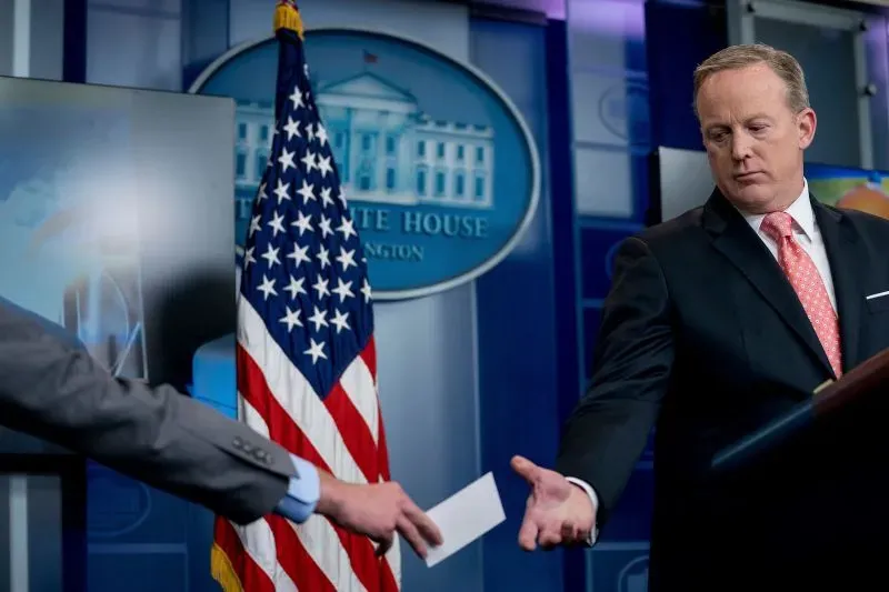 تظهر الصورة شون سبايسر، السكرتير الصحفي السابق، وهو يتلقى بطاقة من صحفي خلال إحاطة في البيت الأبيض، مع العلم الأمريكي في الخلفية.