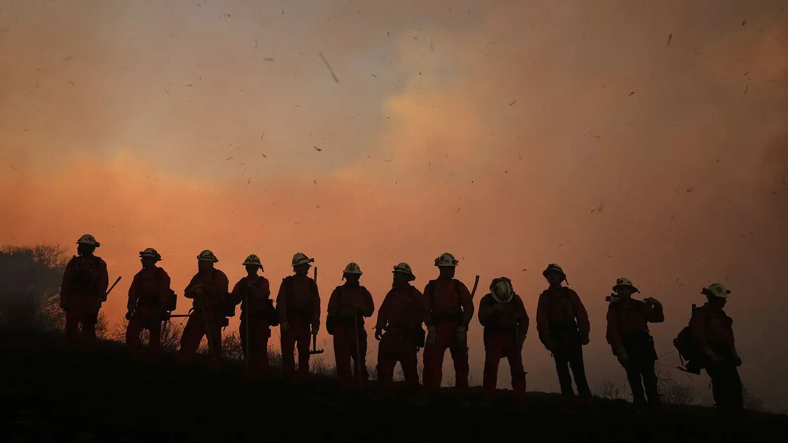 رجال الإطفاء يقفون في صف silhouetted ضد سماء مشتعلة خلال حريق كانيون فاير في شمال لوس أنجلوس، مع تصاعد الدخان واللهب.