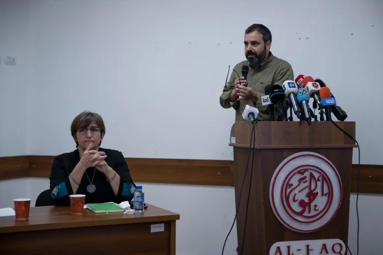 محامٍ يتحدث في مؤتمر صحفي حول حقوق الإنسان في فلسطين، مع وجود صحفيين وأعلام خلفه، يعكس جهود التوثيق والمقاومة.
