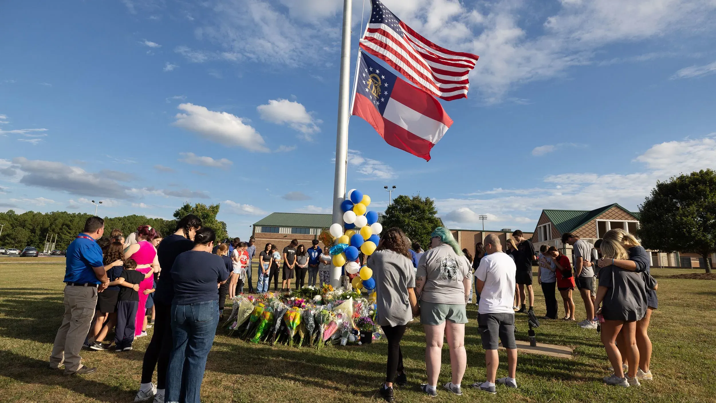 مجموعة من الأشخاص يتجمعون في ساحة مدرسة أبالاتشي الثانوية في جورجيا، أمام نصب تذكاري مزين بالزهور، مع العلمين الأمريكي والجورجي في الخلفية.