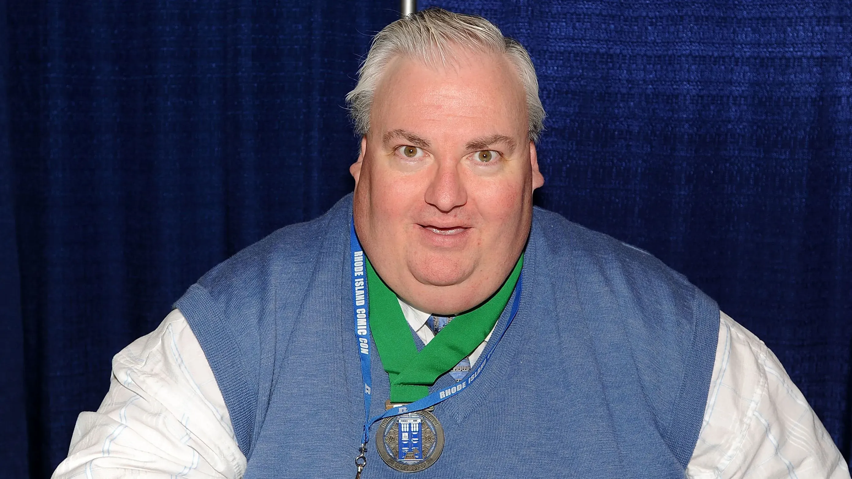 تظهر الصورة سايمون فيشر-بيكر، الممثل البريطاني المعروف بأدواره في "هاري بوتر" و"دكتور هو"، مبتسمًا في حدث جماهيري.