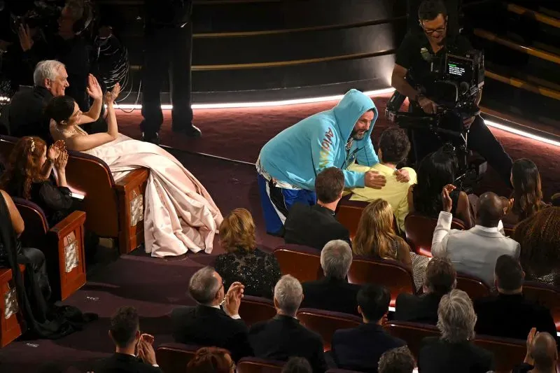 آدم ساندلر يرتدي سترة ملونة وقبعة، محاطًا بالجمهور في حفل الأوسكار، حيث يتفاعل مع أحد الحضور وسط تصفيق الحضور.