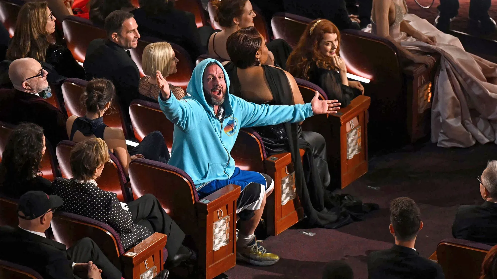 آدم ساندلر يرتدي سترة زاهية وقبعة مع شورت رياضي في حفل الأوسكار، معبرًا عن سعادته وسط جمهور متنوع.