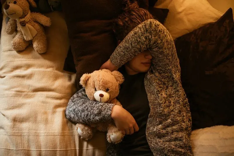 شخص يحتضن دمية دب محشوة أثناء الاستلقاء على السرير، مما يعكس الراحة والتواصل العاطفي المرتبط بالحيوانات المحنطة.