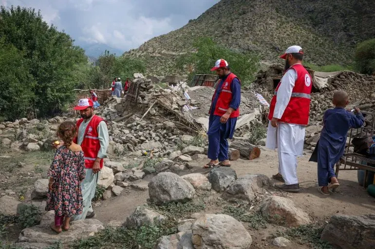 رجال إنقاذ يرتدون سترات حمراء يتفقدون موقع زلزال مدمر في أفغانستان، حيث تظهر الأنقاض والأطفال في الخلفية.