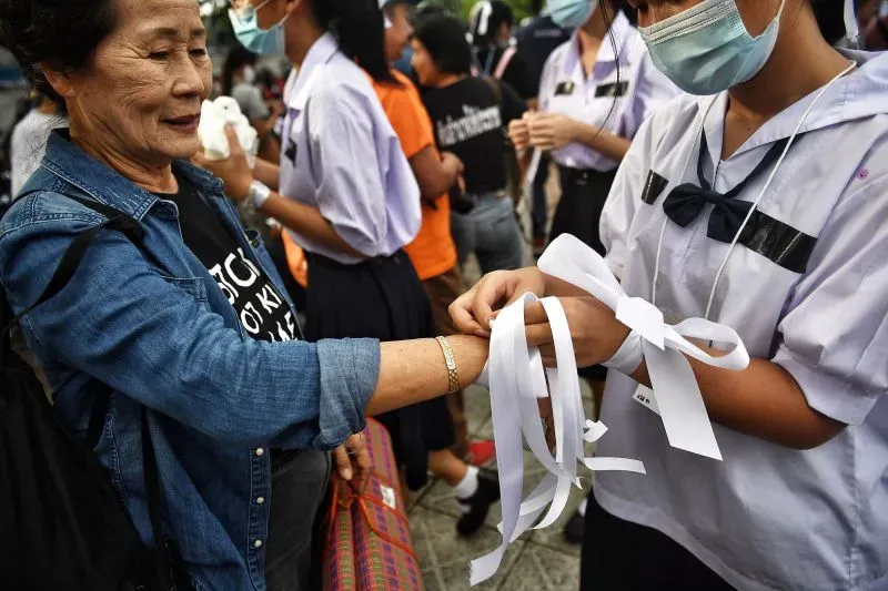 طالب يرتدي زيًا مدرسيًا يربط شريطًا أبيض على معصم امرأة مسنّة، في تجمع طلابي في بانكوك، يعكس التغيير في ثقافة اللباس المدرسي.