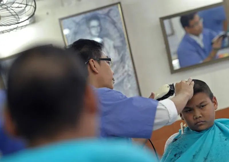 شاب يجلس في صالون حلاقة بينما يقوم الحلاق بقص شعره، مع تعبير جاد على وجهه، مما يعكس الضغوط المرتبطة بقواعد المظهر في المدارس التايلاندية.