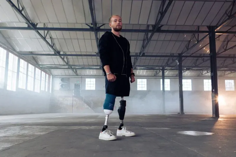 شاب مبتور الساقين يقف في مساحة داخلية، يعكس قوة الإرادة والتصميم بعد تحدياته في سباق الرجل الحديدي.