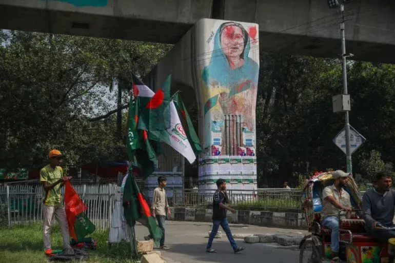 جدارية كبيرة لشيخة حسينة تحت جسر في دكا، مع وجود رجال يحملون أعلام بنغلاديش، تعكس الأجواء السياسية المتوترة في البلاد.