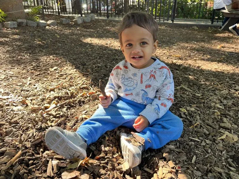 طفل صغير يجلس على الأرض في حديقة، مبتسمًا وهو يحمل عودًا. يرتدي قميصًا مزخرفًا وسروالًا أزرق، محاطًا بأرضية خشبية.