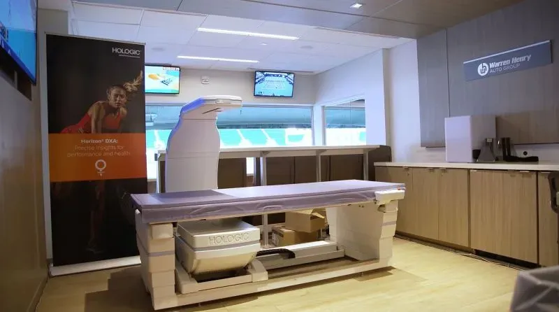 غرفة فحص صحية للاعبات التنس تحتوي على جهاز طبي من شركة Hologic، مع لافتة توضح أهمية الفحوصات الصحية.