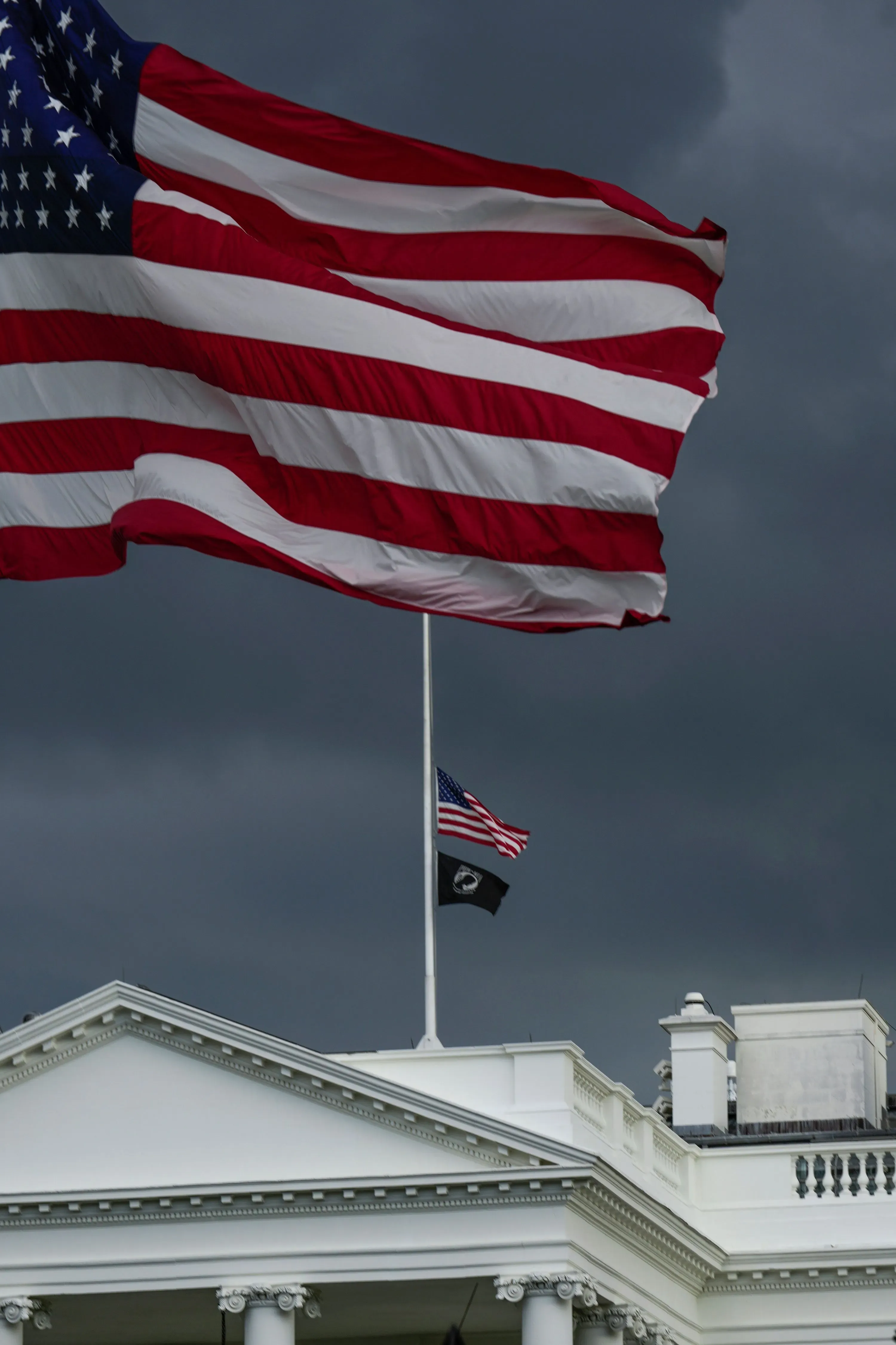 علم الولايات المتحدة يرفرف فوق البيت الأبيض، مع العلم الأسود في الأسفل، في مشهد يعكس التوتر السياسي الحالي.