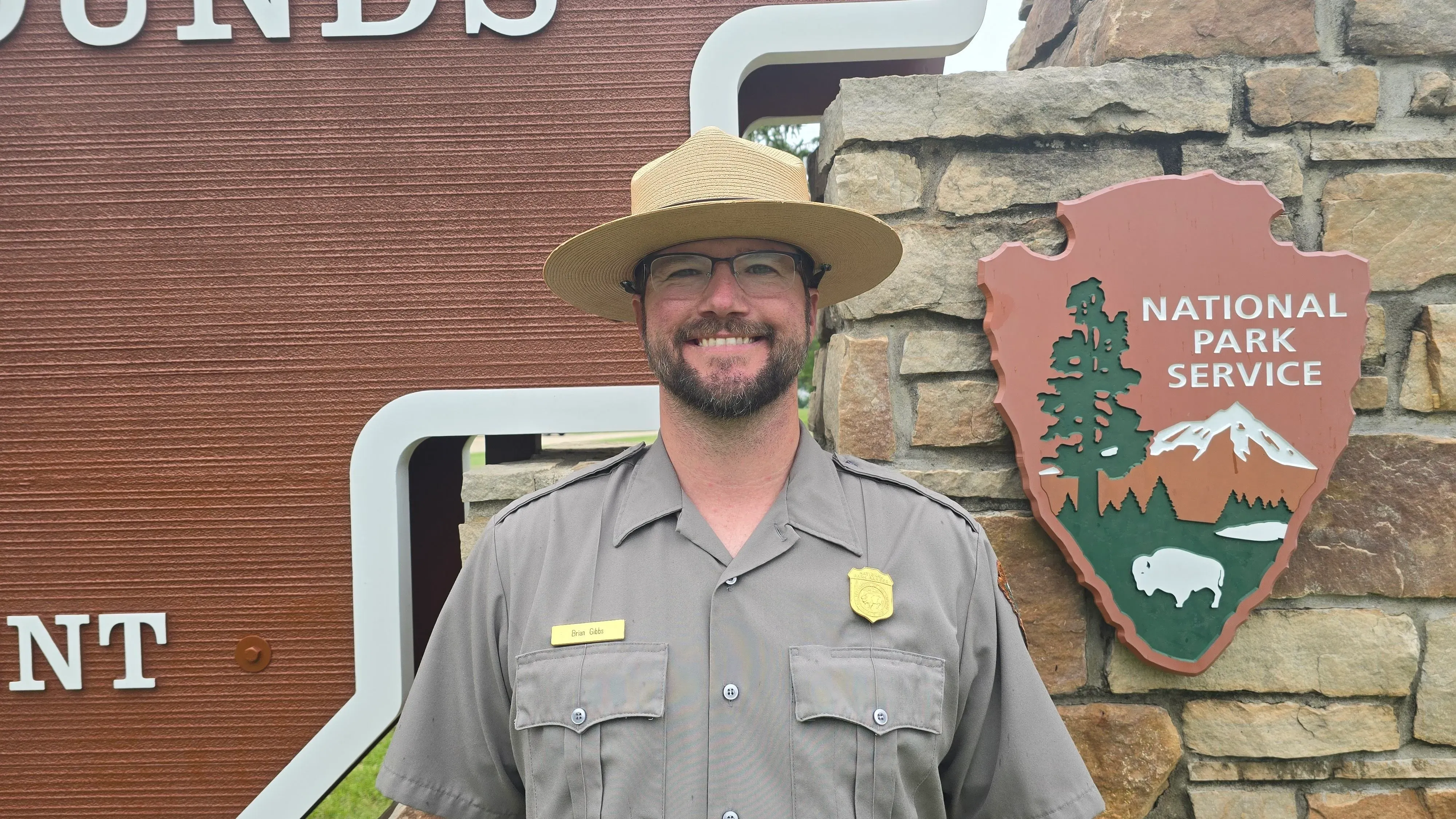 براين غيبس يرتدي زي حارس الحديقة أمام لافتة النصب التذكاري الوطني لتلال إفيجي، حيث كان يعمل على تعليم الزوار عن تاريخ الموقع.