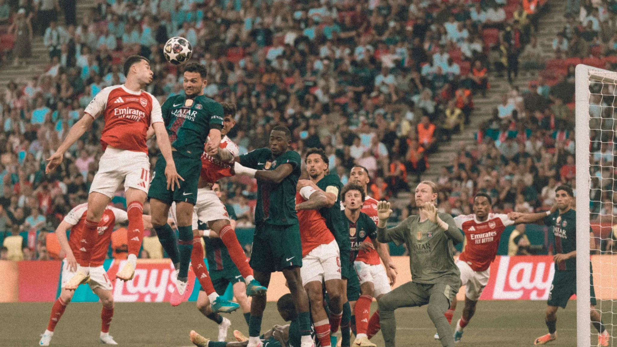 مباراة كرة قدم بين Arsenal وBarcelona تظهر لاعباً من أرسنال يقفز لمنافسة على الكرة في منطقة الجزاء وسط حشد جماهيري كبير.