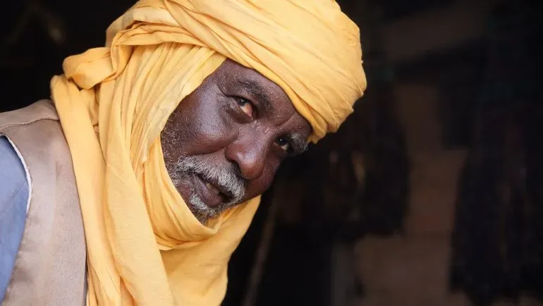 رجل مسن يرتدي عمامة صفراء زاهية، يجلس في متجره لبيع التذكارات في أغاديز، مع تفاصيل عن السكاكين المزخرفة حوله.