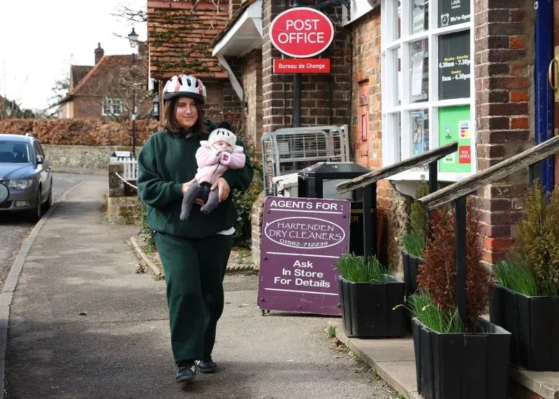 امرأة ترتدي خوذة تحمل دمية في يدها، تسير بجوار مكتب البريد في قرية إنجليزية، وسط شعور بالقلق بسبب هجمات صقر محلي.