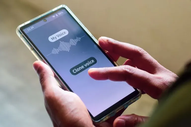 شخص يستخدم هاتفه الذكي لتشغيل تطبيق يتيح له تقليد الأصوات، مع خيارات "صوتي" و"استنساخ الصوت"، مما يعكس تقدم تكنولوجيا الذكاء الاصطناعي.