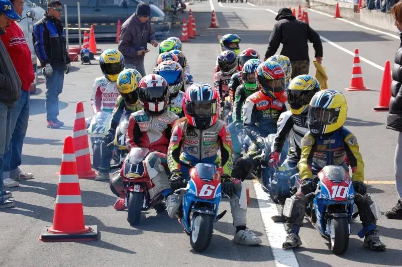 مجموعة من الأطفال يرتدون خوذات ملونة، يستعدون لسباق دراجات نارية صغيرة في حلبة سباق، مع وجود مخاريط مرورية في الخلفية.