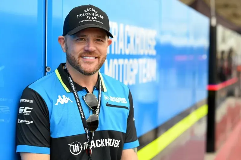 مدير فريق "تراكهاوس" في سباقات الموتو جي بي، مبتسمًا أمام حافلة الفريق الزرقاء، مع شعارات الفريق في الخلفية.