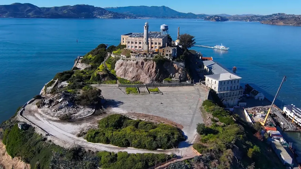 جزيرة الكاتراز الشهيرة في سان فرانسيسكو، تظهر المباني التاريخية المحاطة بالمياه، مع مناظر طبيعية خضراء وجبلية في الخلفية.